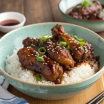 A bowl of tender, Asian-style braised chicken necks glazed in a rich, savory sauce, served over steamed white rice. The dish is garnished with sesame seeds and sliced green onions, with an extra bowl of sauce in the background, enhancing the warm and comforting presentation.